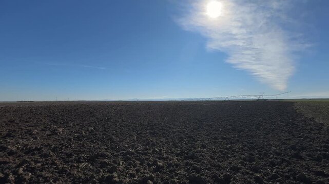 Handheld slow motion shot of dark plowed fields backlit by sunlight, showing a straight horizon and irrigation line under a blue sky with thin clouds.