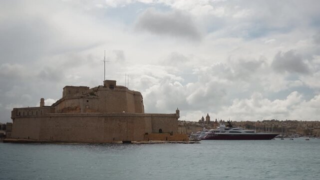 View from passing boat along coastal walls of Fort St Elmo in Valletta. Overcast but warm weather. Massive limestone fortification rises above sea, showcasing its historic military architecture
