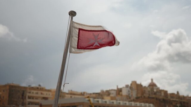 Close-up slow motion of Malta civil ensign flag waving in strong wind on a tourist boat, bright sunlight, daylight. Valletta harbor, yachts and ships, historic architecture blurred in background