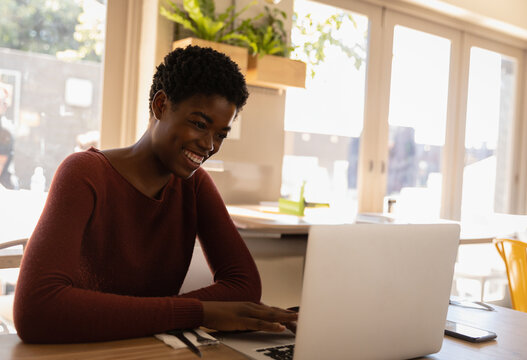 African American woman in twenties wearing maroon sweater sitting at wooden table typing on laptop