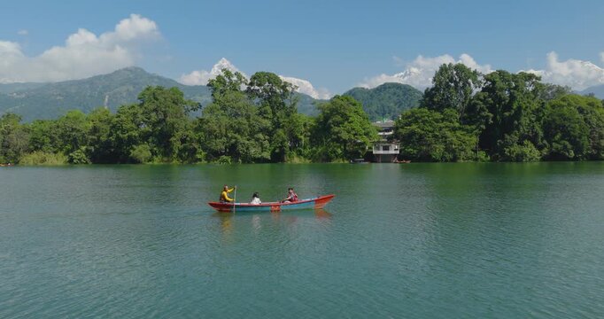 Aerial shot of three villagers rowing a boat across Phewa Lake Nepal Pokhara toward forest and village with Annapurna peaks rising under a cloudy blue sky in Pokhara Nepal.