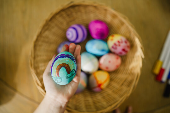 Hand holding painted Easter egg over basket of colorful eggs indoors