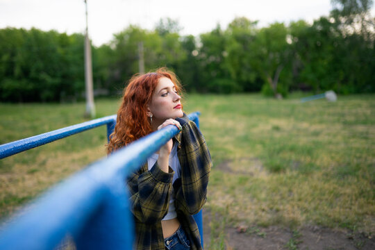 Red haired woman leaning on blue parallel bar outdoors in old playground