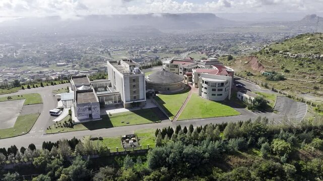 Drone orbits to the left around front of parliament building atop a hill overlooking city on a sunny morning in Maseru, Lesotho