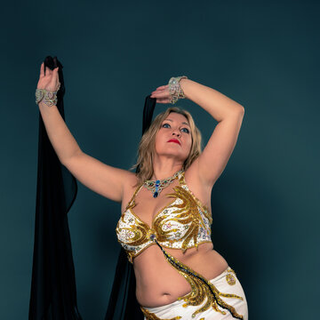 Graceful middle aged woman performs belly dance in an ornate white and golden costume with jewels. Dancer holds black veil against teal studio background. Evokes culture, art and performance