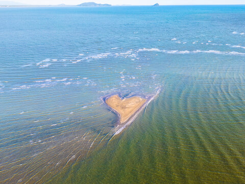 Heart-shaped sandbar floats in calm blue ocean waters. Shimmering ripples reflect the sun's gentle glow. Surrounding sea stretches endlessly to distant horizons