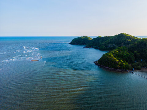 Calm blue waters stretch to distant horizon. Lush green hills rise gently from the sea. Lone sailboat glides softly across the surface. Sunlight bathes the scene with warm, peaceful glow