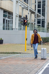 African-American man walking in mustard jacket pulling suitcase using phone by signal, copy space © wavebreak3