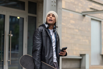 Man leaning against column at entrance wearing beanie holding skateboard checking smartphone © wavebreak3