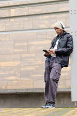 Adult man with beanie leaning against column by tiled wall, checking phone, holding cup, copy space © wavebreak3