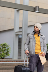Adult male traveler standing on paved plaza by facade holding pastry pulling suitcase, copy space © wavebreak3