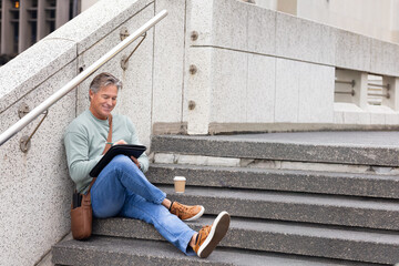 Mature man sitting on urban steps sketching on tablet with stylus, coffee cup, bag, copy space © wavebreak3