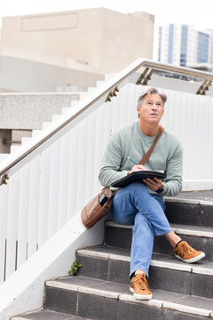 Middle-aged man sitting on outdoor concrete steps holding sketchbook and pen with satchel nearby