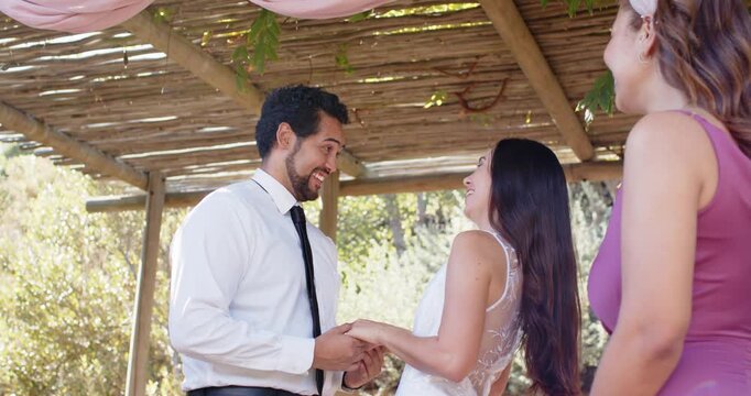 Diverse couple exchanging vows, groom speaking, bride laughing and holding hands under pergola