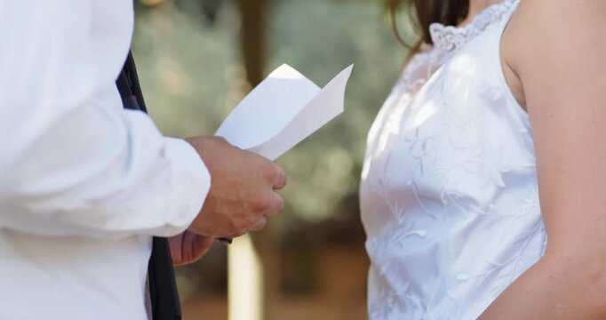 Diverse couple under pergola, groom reading vows from folded sheet, bride listening, ring visible