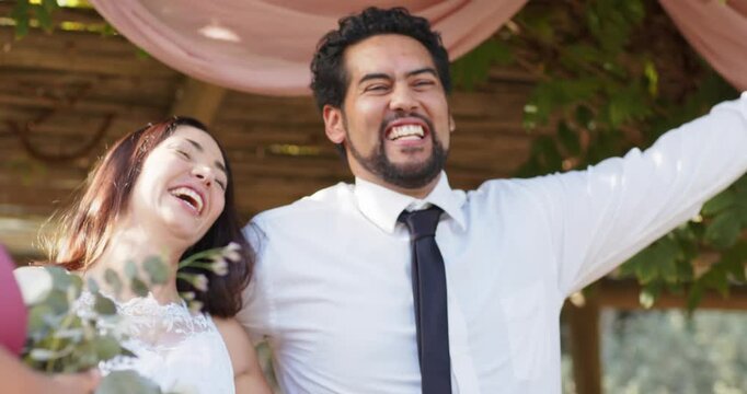 Diverse couple under arch laughing as confetti falls bride in gown with bouquet man greeting guests