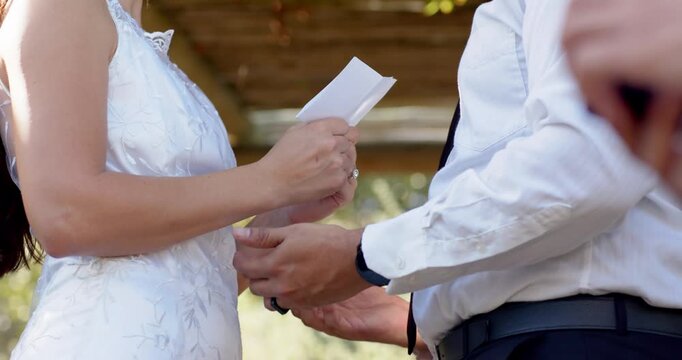 Vertical video: During wedding bride reading folded vows groom reaching clasping hands under arbor
