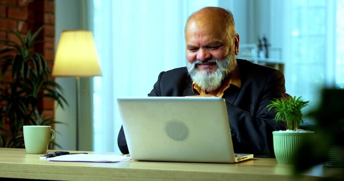 Indian senior businessman or corporate executive listening to a podcast or educational video presentation while working on a laptop in a cozy office environment with professional moody lighting