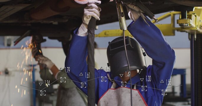 Welding tech wearing helmet, coveralls and apron holding leads in service bay, with grinder sparks