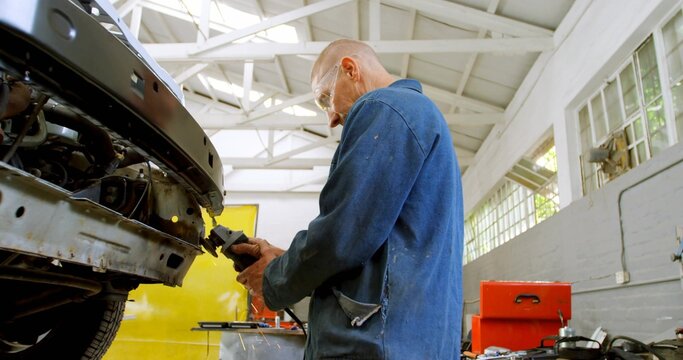 Grinding tech in blue coveralls with goggles, repairing bumperless car front in garage with toolbox