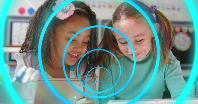 Two girls leaning over tablets tapping in classroom, generating teal rings over them for learning