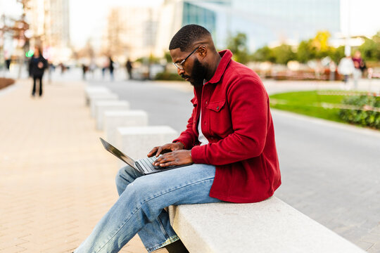 Man in red jacket working on laptop outdoors in urban setting