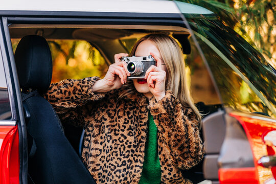 Stylish woman in leopard print coat takes photo from retro car window