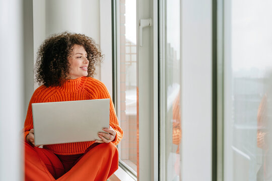 Smiling woman with curly hair using laptop on windowsill indoors