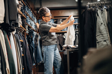 Fototapeta premium Middle-aged Asian woman in gray sweater examines white shirt while shopping in clothing store, surrounded by racks of jeans and apparel in a retail environment