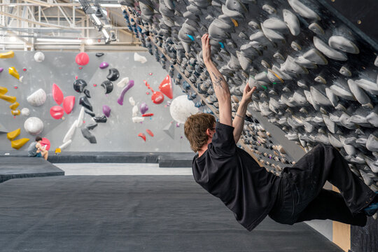 Landscape shot of slim white male with light brown hair mullet haircut, tattoo on right forearm and black frame glasses on indoor rock climbing bouldering overhanging training board with black clothes