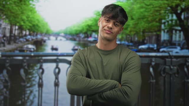 Young man with arms crossed on a canal bridge street, leaning against railing, olive shirt, slight smirk and head tilt; relaxed confidence.