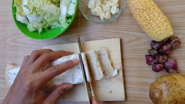 Close up of Hand slices tempeh on wooden board. Cabbage, corn, and potatoes nearby. Fresh ingredients for healthy meal prep. Simple kitchen scene, calm and focused. Perfect for food, health, or cookin