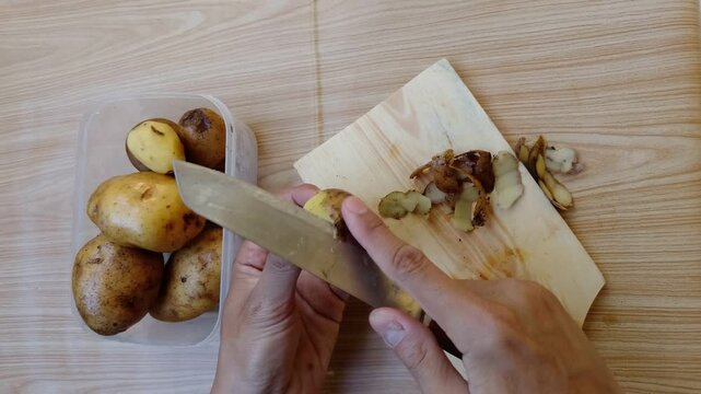 Close up off Hands peel potatoes on a wooden board. Wooden surface shows natural grain texture. Simple kitchen action captured in bright light. No emotion expressed - calm, focused task