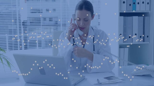 Mid-adult female doctor on phone call, typing updates on laptop, using handset, lab coat in clinic