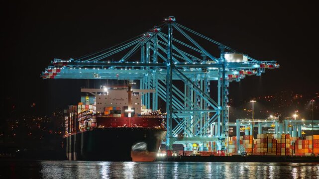 Large quay cranes offloading container from a Suezmax container ship in a bustling port illuminated by night lights. Trucks moving the containers in motion blur.