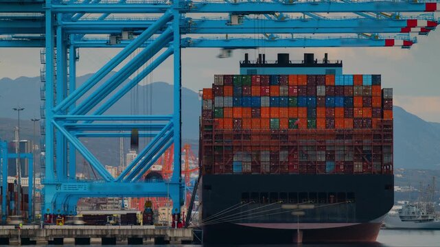 Timelapse of quay cranes offloading containers from a large cargo ship onto semi-trucks moving in motion blur. Mountains and moving clouds in the background. Busy port operations on a cloudy day.