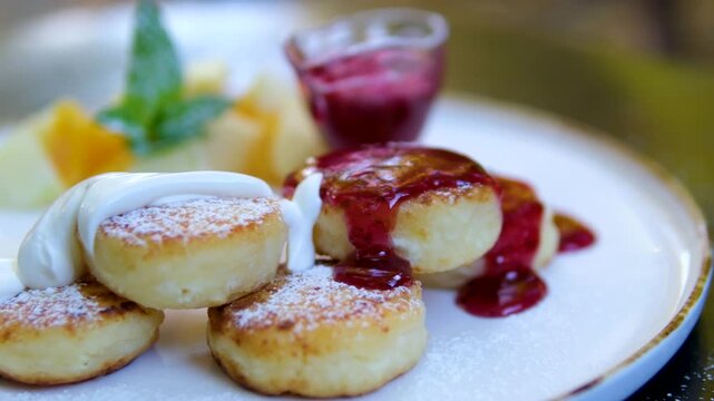 Cottage cheese pancakes served on white plate with jam, sour cream, fruit pieces and mint. Powdered sugar is sprinkled and sour cream is poured. Close-up and top view of a cozy dessert breakfast set.