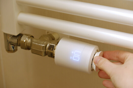 Modern smart thermostat head on white towel radiator in bathroom, white LCD screen showing temperature settings, hand adjusting dial. Energy saving, and modern climate control.