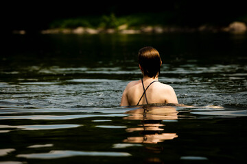 Fototapeta premium Woman from behind swimming in a dark natural lake or river, her wet hair and swimsuit strap visible above the water in a quiet and contemplative outdoor swimming scene.
