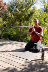 African-American man kneeling on yoga mat on deck, in red, water bottle by railing, copy space © wavebreak3