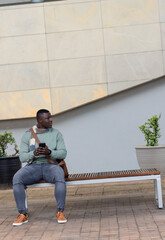 African American man sitting on wooden bench at brick plaza holding smartphone and crossbody bag © wavebreak3