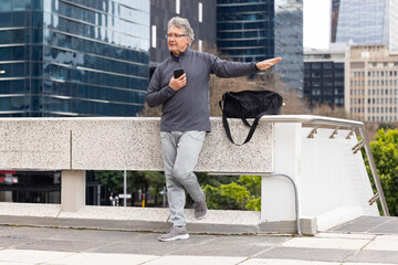 Senior man standing on elevated urban terrace, holding smartphone and black duffel bag, gesturing © wavebreak3