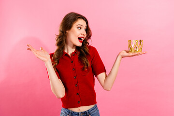Young chic woman in red top holds a small gold crown smiling against a pink studio backdrop for a fashion beauty and glamour themed stock image