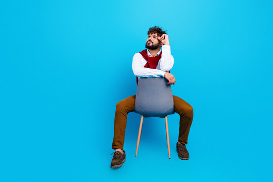 Thoughtful young man sitting backward on a chair against a bright blue background wearing casual smart clothes and sweater draped over shoulders