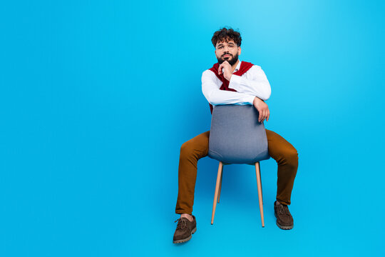 Casual young man sitting backwards on a chair against a bright blue background, looking confident and thoughtful in modern fashion
