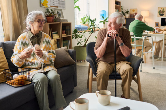 Senior Caucasian woman knitting while senior Caucasian man resting on cane and conversing in care home lounge. Supporting retirement living, elder care, community service marketing