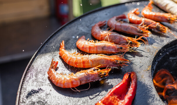 Variety of food being cooked on a grill. The food includes shrimp, corn on the cob, and flatbreads, which are all being cooked over an open fire. The food is being cooked in a round metal pan.
