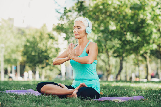 Gentle senior woman practices yoga in the park outdoor fitness moment with headphones for wellness and balance in nature