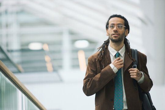 Portrait of young adult man adjusting tie and carrying bag while walking through modern office building, representing professional confidence. Ideal for career growth, recruitment, corporate branding