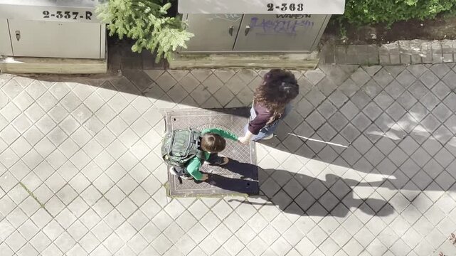 Madre con hijo en vuelta al cole de la mano vistos desde arriba y sombra alargada en acera de calle